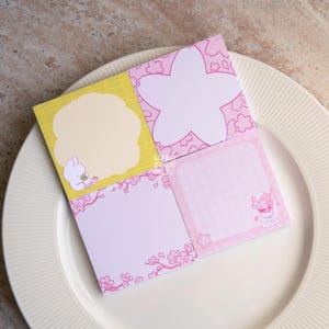Op de afbeelding: Een vierkant notitieblok met vier secties, elk met een ander ontwerp. Eén sectie heeft een gele achtergrond met een konijnenillustratie. De andere secties hebben roze kersenbloesem- en dessertillustraties. Het notitieblok ligt op een wit bord.