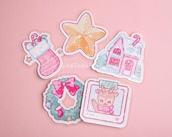 May include: A collection of Christmas-themed stickers. The stickers include a pink and white stocking, a gold star, a pink house with snow, a wreath with a pink bow, and a deer wearing a scarf. The stickers are on a pink background.