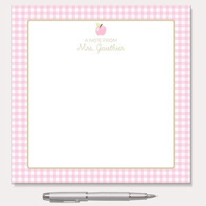 Peut inclure: Un bloc-notes à carreaux roses et blancs avec une bordure dorée et le texte "A note from Mrs. Gauthier" et une petite illustration de pomme. Un stylo argenté repose en dessous.