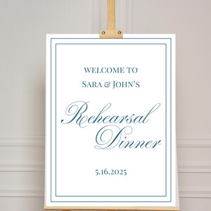 Könnte beinhalten: Ein weißes Schild mit blauem Rand und dem Text "WELCOME TO SARA & JOHN'S Rehearsal Dinner 5.16.2025" wird auf einer hölzernen Staffelei angezeigt. Der Text ist in dunkler blauer Schrift.