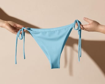 Könnte beinhalten: Hellblaue Bikinihose mit Bindebändern an den Seiten. Die Bademode wird von zwei Händen vor einem neutralen Hintergrund gehalten. Die Bikinihose hat eine klassische Dreiecksform.