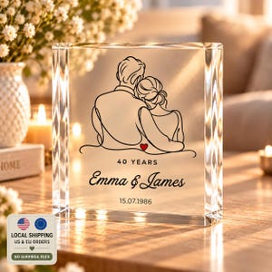 Op de afbeelding: Een rechthoekig, helder glazen blok met een zwarte lijntekening van een omhelzend stel met een klein rood hartje. De tekst luidt 40 YEARS, Emma & James en 15.07.1986. Het glas heeft afgeschuinde randen en een gladde, transparante afwerking.