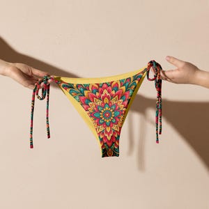 Könnte beinhalten: Gelber Bikini-Slip mit einem lebendigen Blumenmuster in Rot, Türkis und Gold. Der Bikini hat Bindebänder mit passenden gemusterten Schnüren. Das Kleidungsstück wird von zwei Händen vor einem neutralen Hintergrund gehalten.