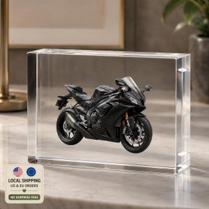 Puede incluir: Un modelo de motocicleta deportiva negra encapsulado dentro de un bloque acrílico rectangular transparente. El vehículo en miniatura presenta ruedas detalladas, un parabrisas y un tubo de escape lateral, suspendidos en el material.