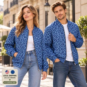 May include: Two matching blue bomber jackets featuring a dark blue leopard print pattern. Each jacket has a full-length front zipper, long sleeves, and a ribbed collar. The jackets are worn over plain white t-shirts and blue denim jeans.