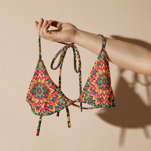 May include: A colorful bikini top with a floral pattern in shades of red, yellow, and teal. The triangle top has tie straps and a matching design. The garment is held up by a hand against a neutral background.