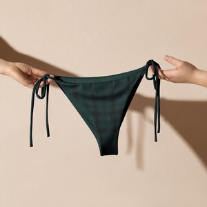 Può includere: Slip bikini verde scuro con motivo a quadri neri. Lo slip bikini ha lacci neri su ogni lato. L'indumento è tenuto da due mani su uno sfondo neutro.