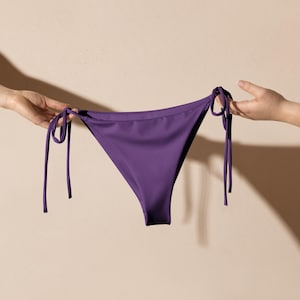 Puede incluir: Braguita de bikini morada lisa con lazos laterales. La prenda de baño está sujeta por dos manos sobre un fondo neutro. La braguita de bikini tiene un corte clásico y está hecha de un tejido liso y brillante.