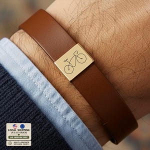 May include: A brown leather bracelet featuring a rectangular gold-toned metal charm engraved with a simple line drawing of a bicycle. The bracelet is worn on a wrist, partially covered by the cuff of a light blue shirt and a dark navy blue jacket.