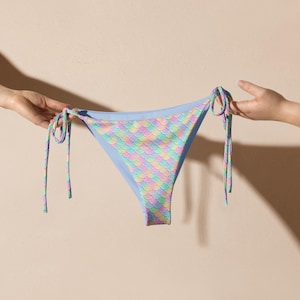 May include: A light blue bikini bottom with a mermaid scale pattern in pastel colors of pink, yellow, and green. The bikini bottom has tie strings on each side.