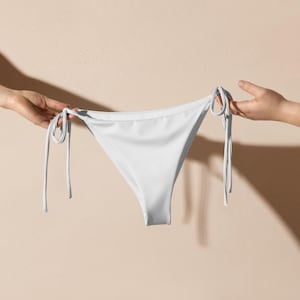 Puede incluir: Braguita de bikini blanca con laterales anudados. La braguita está sujeta por dos manos sobre un fondo neutro. La braguita de bikini es de color blanco liso y tiene una forma triangular clásica.