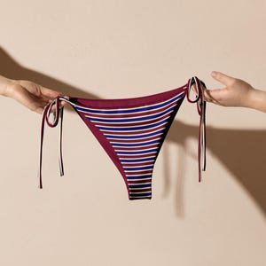 Op de afbeelding: Bordeauxrode en gestreepte bikinibroek met witte, marineblauwe en bordeauxrode horizontale strepen. De bikinibroek heeft aan elke kant strikkoorden. De achtergrond is effen en neutraal.