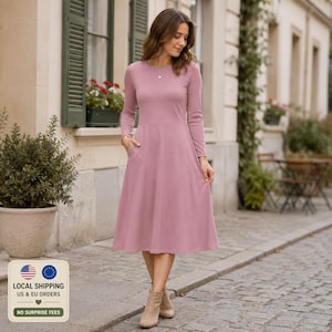 May include: A dusty rose midi dress featuring long sleeves, a round neckline, and a fitted bodice that transitions into a flared skirt. The dress includes functional side pockets and is crafted from a solid-colored, smooth fabric.