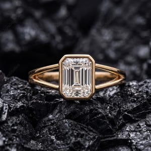 May include: A gold ring with a large, rectangular, clear gemstone. The ring's band splits into two delicate strands on either side of the gemstone. The ring is set against a dark, textured background.