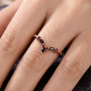 Könnte beinhalten: Ein roségoldener Ring mit V-förmigem Design. Der Ring hat drei schwarze Steine im Marquiseschliff und vier runde rote Steine. Der Ring befindet sich am Finger einer Person.