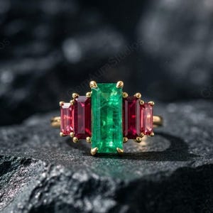 May include: A gold ring featuring a large, rectangular emerald center stone flanked by two smaller, rectangular ruby stones on each side. The gemstones are set in a gold band, creating a striking contrast against the dark background.