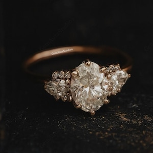 Puede incluir: Un anillo de compromiso de oro rosa con una gran piedra central de diamante de talla ovalada, flanqueada por piedras laterales en forma de pera y diamantes de acento más pequeños. El anillo está sobre un fondo oscuro y texturizado.