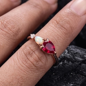 Anello asimmetrico a tre pietre con rubino e opale a forma di pera in oro rosa, gioiello unico con pietra portafortuna di luglio e ottobre, anello di promessa alternativo per la moglie.