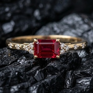 Peut inclure: Une bague en or ornée d'une grande pierre rouge foncé de forme rectangulaire en son centre. De petites pierres claires ornent les côtés de la pierre principale et le long de l'anneau. La bague est posée sur un fond sombre et texturé.