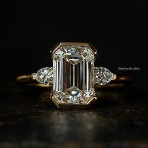 May include: A gold ring with a large, rectangular emerald-cut diamond centre stone, accompanied by two pear-shaped diamonds. The ring is set against a dark backdrop, accentuating the sparkle of the gems. The text "GemAuraStudios" is visible.