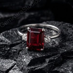 May include: A silver ring with a large, rectangular, deep red gemstone as the focal point. Two smaller, clear gemstones flank the main stone. The ring is set against a dark, textured background, highlighting the jewelry's brilliance.