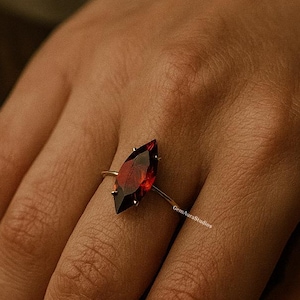 Puede incluir: Un anillo de oro con una gran gema roja oscura con corte marquesa. La gema está engastada en una delicada banda de oro, creando un contraste llamativo. El anillo se muestra en una mano, mostrando su elegante diseño y color vibrante.
