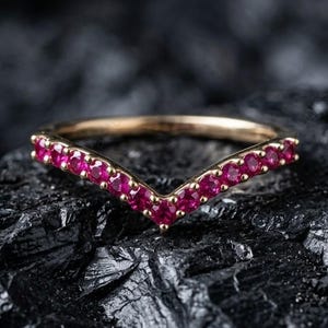 Puede incluir: Un anillo dorado con un diseño en forma de V. El anillo está adornado con una fila de pequeñas y vibrantes piedras preciosas rojas. El anillo se encuentra sobre un fondo oscuro y texturizado.