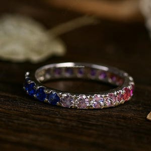 May include: A silver ring with a gradient of colorful gemstones. The ring transitions from blue to pink to red. The ring is set on a dark wooden surface, with a blurred background.