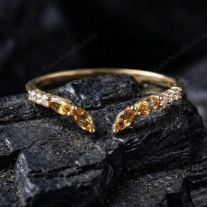 Könnte beinhalten: Ein goldfarbener Ring mit geteiltem Schaft, der drei marquiseförmige, bernsteinfarbene Edelsteine und kleine, klare Akzentsteine aufweist. Der Ring ist vor einem dunklen, strukturierten Hintergrund platziert, der sein elegantes Design hervorhebt.