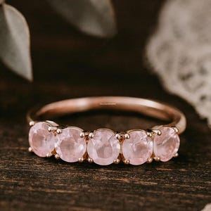 May include: A rose gold ring featuring five round, light pink gemstones. The ring is set on a dark wooden surface, with a soft, blurred background. The gemstones are evenly spaced and appear to be polished.