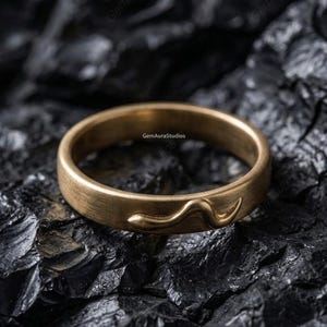 May include: A gold-coloured ring with a brushed finish. The ring features a stylised snake design on the band. The ring is set against a dark, textured background, highlighting the jewellery's details.