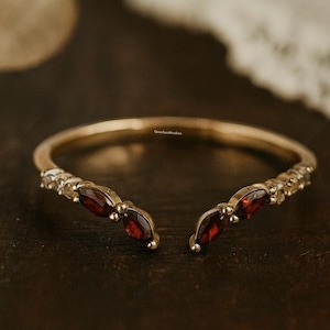 Op de afbeelding: Een goudkleurige open ring met een gespleten ontwerp, met kleine, sprankelende stenen en drie dieprode marquise-geslepen edelstenen aan elk uiteinde. De ring is geplaatst op een donker, getextureerd oppervlak.