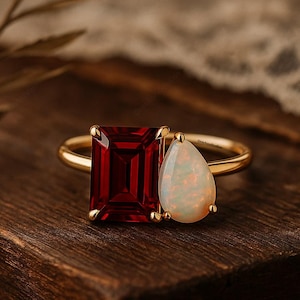 Könnte beinhalten: Ein goldener Ring mit einem großen, rechteckigen, tiefroten Edelstein und einem birnenförmigen, opalisierenden weißen Edelstein. Der Ring liegt auf einer Holzoberfläche, die das Design und den Farbkontrast des Schmucks hervorhebt.