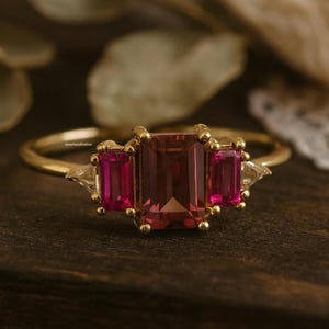 Può includere: Un anello d'oro con una grande pietra centrale rettangolare, marrone rossiccio, affiancata da due pietre più piccole, rettangolari, color magenta. L'anello ha una fascia delicata e piccole pietre di accento su entrambi i lati delle pietre centrali.