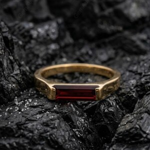 May include: A gold-colored ring with a rectangular, deep red gemstone set in the center. The ring is displayed on a dark, textured surface, creating a contrast between the shiny metal and the rough background. The ring's design is simple and elegant.