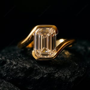 Può includere: Un anello d'oro con un diamante taglio smeraldo. Il diamante è incastonato in una montatura a castone e ha una forma rettangolare. La fascia ha un design liscio e curvo. L'anello è esposto su una superficie scura e strutturata.