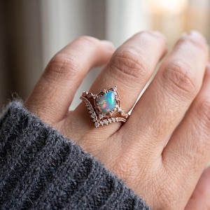 Peut inclure: Une bague en or rose ornée d'une opale en forme de poire aux reflets irisés. L'anneau présente une forme en chevron et est agrémenté de petites pierres claires serties pavé le long des bords et autour du cadre de la monture centrale.