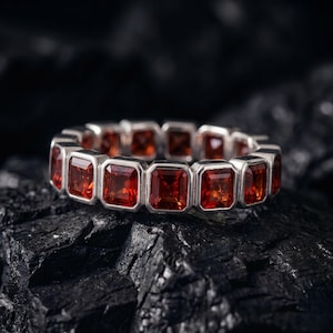 May include: A silver eternity band ring featuring a continuous row of rectangular, faceted, deep orange-red gemstones set in individual metal frames. The ring is positioned horizontally, showcasing the uniform cut and vibrant color of each stone.
