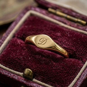 May include: A gold signet ring featuring a polished finish and a smooth, oval-shaped face. The center of the ring is engraved with a single, elegant cursive letter D. The band is solid gold and tapers slightly toward the back of the ring.