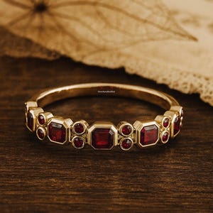 Op de afbeelding: Een gouden bandring met afwisselende rechthoekige en ronde rode edelstenen. De ring ligt op een donkere houten ondergrond, met een vage achtergrond van een gedroogd blad en stof. De ring is een sieraad.