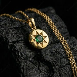 May include: A gold-colored pendant necklace featuring a round pendant with a starburst design and a central emerald-green gemstone. The necklace chain is also gold-colored. The pendant is approximately 0.75 inches in diameter.