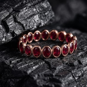 Könnte beinhalten: Ein Ring mit einem durchgehenden Band aus oval geschliffenen, tiefroten Rubinen, die in einen polierten Rahmen aus Roségold gefasst sind. Die Steine sind in einer gleichmäßigen Reihe angeordnet und bilden ein kreisförmiges Design.