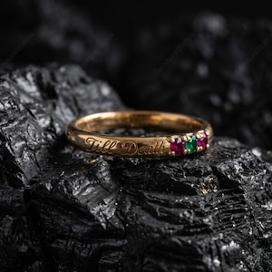 Pode incluir: Um anel de ouro com a inscrição "Till Death" e três pedras preciosas: vermelha, verde e vermelha. O anel é apresentado sobre um fundo escuro e texturizado, realçando sua superfície polida e detalhes intrincados. Uma joia.