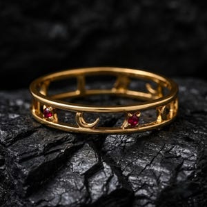 Könnte beinhalten: Ein goldener Bandring mit einer Reihe von ausgeschnittenen Mondsichelformen, die sich mit kleinen, runden, leuchtend roten Edelsteinen abwechseln, welche in den Metallrahmen eingefasst sind und ein durchgehendes durchbrochenes Design bilden.