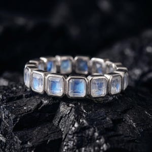 May include: A silver-toned eternity ring featuring a continuous row of square-cut moonstones. Each stone is set in a polished metal bezel, displaying a translucent, iridescent blue glow that shifts across the surface of the faceted gems.