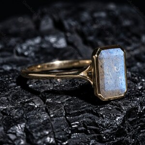 May include: A gold ring with a rectangular, light blue gemstone. The gemstone is set in a gold bezel. The ring is on a dark, textured surface. The ring's band is thin and delicate.