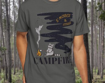 May include: A gray t-shirt featuring a raccoon roasting a marshmallow over a campfire. The shirt has the words "S'mores & Stars" and "Campfire" printed on it in a distressed font. The design includes a dark cloud and stars.