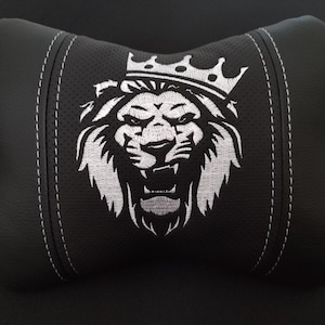 May include: A black bone-shaped car headrest pillow featuring a central panel of perforated material. The pillow is decorated with a white embroidered graphic of a lion wearing a crown. The design includes white stitched seams on both sides.