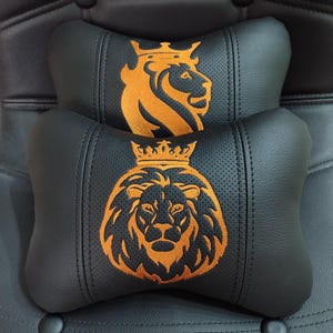 May include: Two black car headrest pillows with an orange lion crest design. The pillows are made of a perforated material and are stacked on top of each other. The lion crest features a crown.