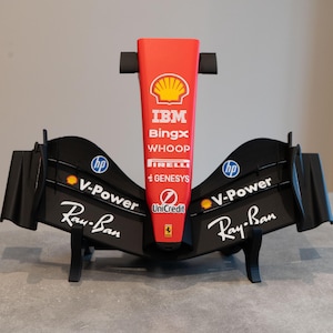 Puede incluir: Una pieza de exhibición de morro y alerón delantero de coche de Fórmula 1 en rojo y negro. La sección central roja presenta logotipos de Shell, IBM, BingX, Whoop, Pirelli, Genesys, UniCredit y Ferrari. Las alas negras muestran logos de HP, V-Power y Ray-Ban.